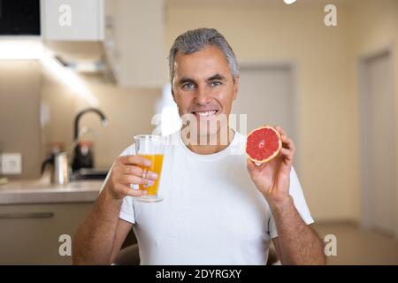 Good loking senior man breakfasts. Vegan food. Mature man in white casual t-shirt holding half of grapefruit and glass of citrus juice and smiling Stock Photo