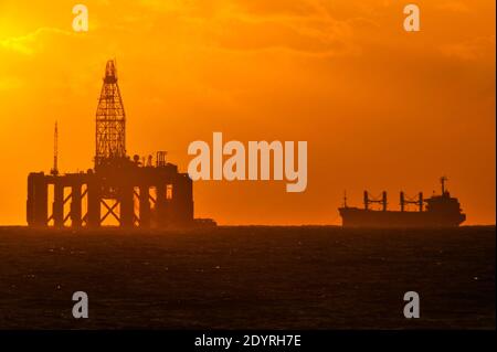 Sun setting over a natural gas drilling platform at sea, with support tanker in the background. Stock Photo