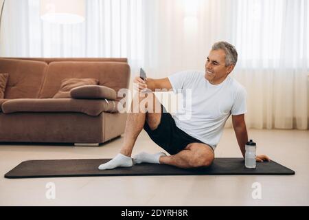 Procrastination during fitness. Senior man resting after home workout with water and phone. Sporty man after practicing yoga, break in doing exercise Stock Photo