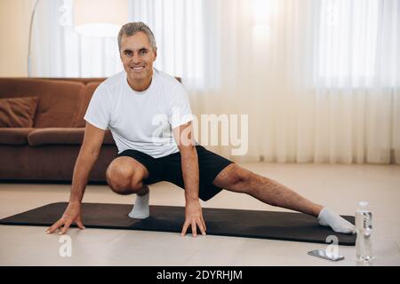 Elderly man doing side lunge exercise. Senior male athlete with fit body warming up, doing leg stretch workout at home. Take care of yourself Modern Stock Photo