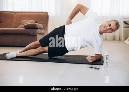 Cheerful senior man doing side plank exercise on yoga mat. How to stay healthy on quarantine concept. Healthy lifestyle. Athletic man training and Stock Photo