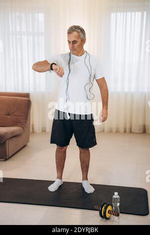 Fit man monitors his heart rate with a smart watch. Workout at home concept. Man with blue jumping rope around neck, standing on yoga mat and looking Stock Photo