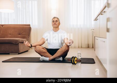 Sporty mature man meditating after yoga practice. Man sitting on black yoga mat with eyes closed in modern living room and relaxing in easy seat pose Stock Photo