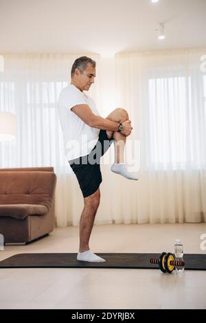 Mature man standing on one leg and stretching another. Workout on black yoga mat. Fit man practicing yoga position at home. Morning warm up. Light Stock Photo
