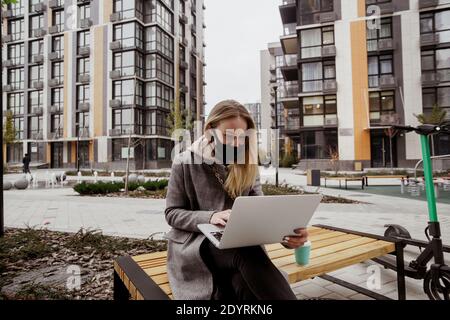 Blonde woman came to this park on electric scooter. She sitting on bench, using her modern laptop and talking by video call with her friends. Black Stock Photo