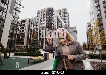 Posing for photo Young cheerful blonde woman showing peace sign and looking at camera with smile. Grey modern apartment blocks on background. She Stock Photo