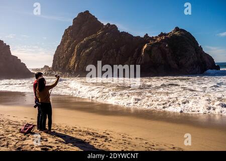 Asian couple taking a selfie in front of iconic sea cave at Pfeiffer Beach in Big Sur, California, USA Stock Photo