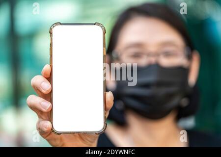 Blurred Asian middle-aged woman showing smartphone, blank space for copy and design on smart phone, woman wearing black medical mask to protect hersel Stock Photo