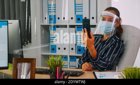 Entrepreneur wearing face protection against coronavirus using phone for videomeeting talking with remotely coworker sitting in new normal office corporate building. Woman respecting social distance Stock Photo