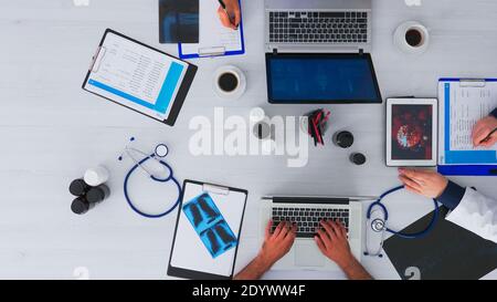 Top view of doctors discussing about virus using tablet, taking notes sitting on desk in medical clinic with digital technology. Assistants writing on laptop and clipboard, researching on copy space Stock Photo