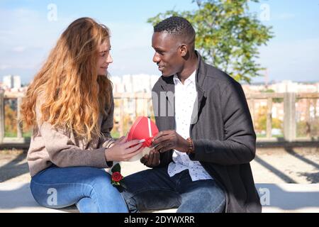 African black man and redhead caucasian woman with a rose opening a heart shape box with sweets and looking each other. Young multiracial couple celeb Stock Photo