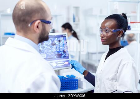 African scientist discussing result of virus research with supervior in laboratory using computer. Multiethnic team of medical researchers working together in sterile lab wearing protection glasses and gloves. Stock Photo