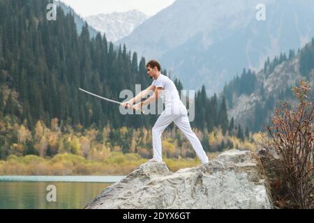 A young man stands on a large stone and holds a Japanese sword in his hands. Stock Photo