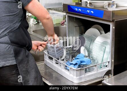 Restaurant kitchen worker emptying a dishwasher removing clean cutlery from the basket in a close up on his hands and the open door of the machine Stock Photo