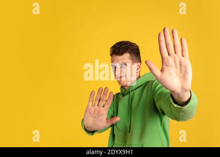 Stop, rejecting. Caucasian young man's portrait on yellow studio background. Beautiful male model in green outfit gesturing. Concept of human emotions, facial expression, sales, ad, youth. Copyspace. Stock Photo