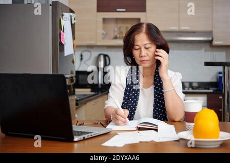 Serious senior woman sitting at kitchen table, talking on phone and taking notes in planner Stock Photo
