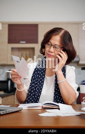Portrait of serious senior woman talking on phone and checking utility bills when sitting at kitchen table Stock Photo