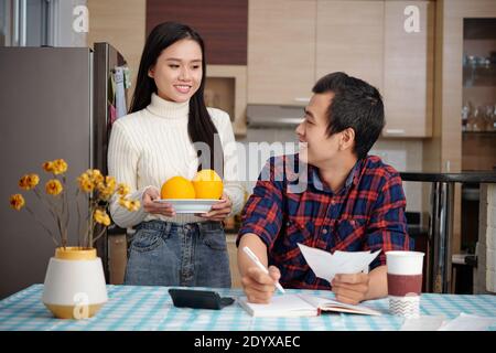 Handsome smiling Vietnamese man checking bills and writing down expenses and incomes when girlfriend briging him plate with two oranges Stock Photo