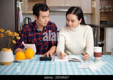 Young Asian couple reviewing bills, planning family budget and calculating finances at kitchen table with bills, calculator and planner Stock Photo