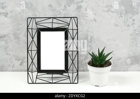 Black metal vertical frame with white blank and succulent flower on table against grey concrete wall. Mockup Template for your design, text. Stock Photo