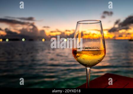 Glass of white wine on a table with dramatic sunset in the background and lights on horizon, Maldives. Stock Photo