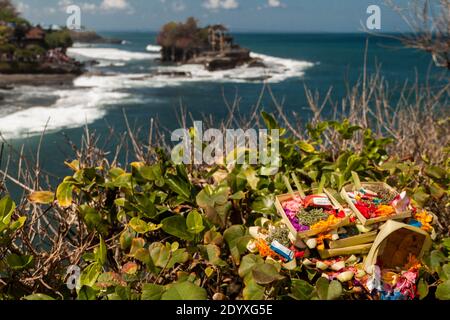 Canang sari daily offerings placed on the bushes by the wavy sea and Tanah Lot Temple in the blurry background Stock Photo