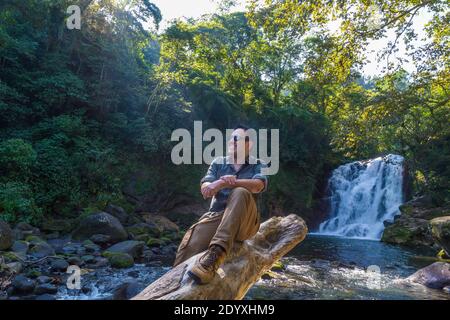 Handsome Hispanic man sitting on a stone with a flowing waterfall behind  and smiling happily Stock Photo