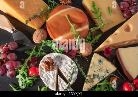 cheese background. Assortment different types cheese with walnuts, tomato and grapes on black slate background. blue cheese Dorblu, smoked, brie, Maas Stock Photo