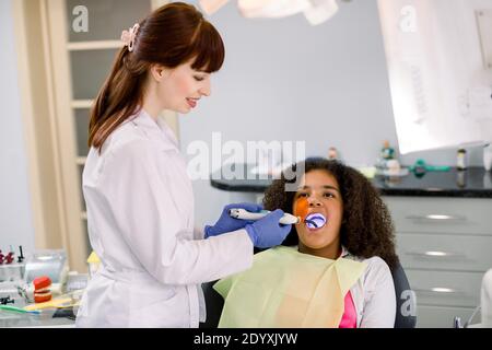 Female dentist performing dental filling procedure to a little mixed raced school girl in pediatric dental clinic. Doctor using dental ultraviolet Stock Photo