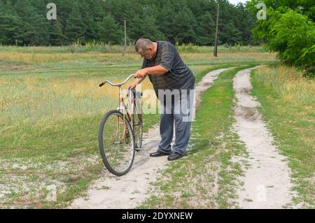 Senior Ukrainian peasant standing on a country road and inspecting old rusty bicycle with broken saddle Stock Photo
