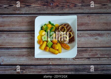Beef steak with potato croquettes and vegetables (green asparagus and yellow carrots) on a white square plate with dark wooden background. Stock Photo