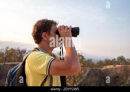 A young man with a backpack on his back watches through binoculars from the height of the mountains. Stock Photo
