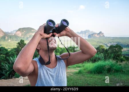 A young man looks into the sky through binoculars. A traveler on the background of wild nature. Stock Photo