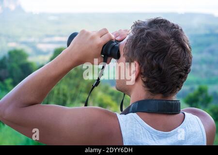 A young man looks through binoculars from the top on the background of the forest. Stock Photo