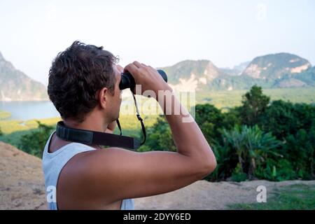 Young male traveler looks through binoculars from the top against the backdrop of rocks and tropical forest. Stock Photo