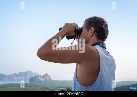 A young man in a white T-shirt looks through binoculars from the top against the background of the sky and rocks. Stock Photo