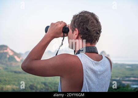A young man in a white T-shirt looks from the top into the distance through binoculars at the wildlife. Stock Photo