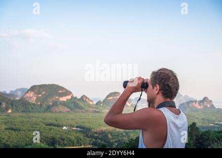 A young man in a white T-shirt looks from the top into the distance through binoculars against the backdrop of wildlife, sky, mountains and sea. Stock Photo