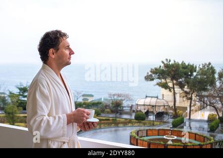 A man in a white bathrobe meets the morning with a cup of tea or coffee overlooking the sea. The concept of relaxation and health. Stock Photo