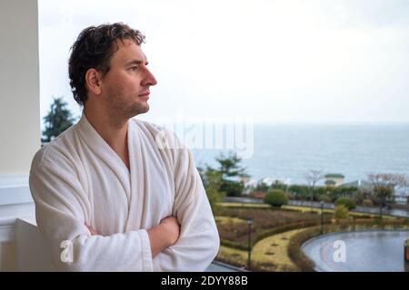 Close-up portrait of a man in a white bathrobe who is standing on the balcony of the hotel overlooking the sea. Stock Photo