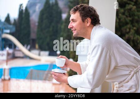 Closeup portrait of a cheerful happy man in a white bathrobe and with a cup of morning tea or coffee. Stock Photo