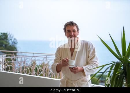 A smiling Caucasian man in a white bathrobe with a cup of tea or coffee in his hands looks at the camera. He enjoys a fresh invigorating morning overl Stock Photo