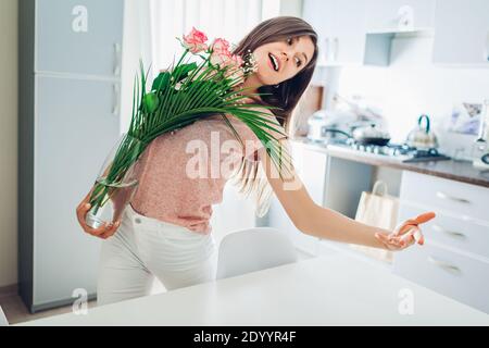 Happy woman puts vase with flowers dancing and singing. Girl taking care of coziness on kitchen on quarantine isolation at home. Stock Photo
