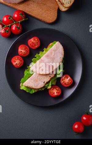 Sandwich with turkey ham meat, green salad and fresh cherry tomatoes slices on black plate near to ingredients on cutting board, grey minimal backgrou Stock Photo