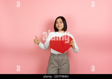 close up a joyful cheerful Asian woman holds a large red paper heart in her hands and gives a thumbs up isolated on a pink background Stock Photo