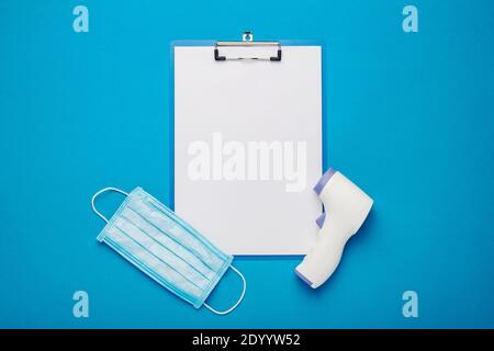 Digital non-contact thermometer, face shield and clipboard on a blue background. Banner. Top view, flat lay. Stock Photo