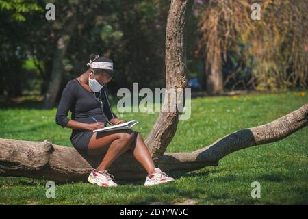 Woman with a cloth face mask sitting on a lying tree trunk writing in a questionnaire before passing a test against the corona virus. Stock Photo