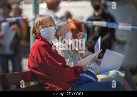 Three senior women wearing facial masks sitting on a bench next to a queue of people waiting for a corona virus test. Stock Photo