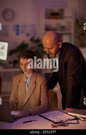 Mature finance manager helping his young female colleague at work checking data on laptop late in evening Stock Photo