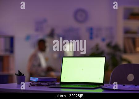 Modern office workspace late in evening with laptop on foreground and unrecognizable man working on background, copy space Stock Photo